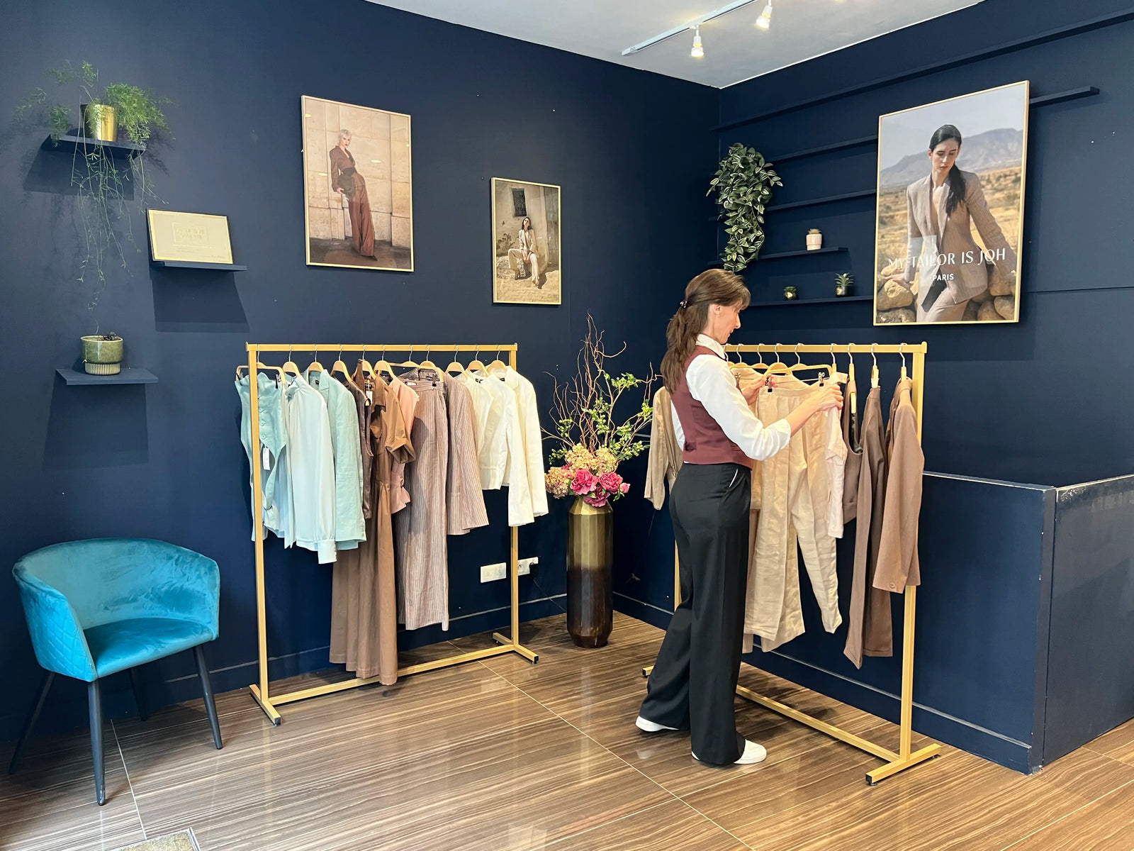 Intérieur de la boutique My Tailor Is Joh à Paris : conseillère en style présentant une collection de tailleurs et chemisiers femme dans un showroom aux murs bleu nuit et portants dorés.