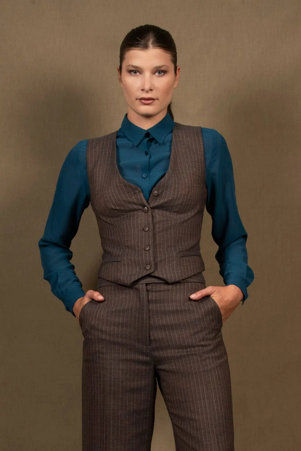 Gilet tailleur femme en 100% laine, couleur marron rayé. Cinq boutons en nacre mates ton sur ton sur le devant, ainsi que deux poches passepoilées. La coupe de ce gilet est cintrée. La doublure est en 100% viscose avec un coloris ambré.