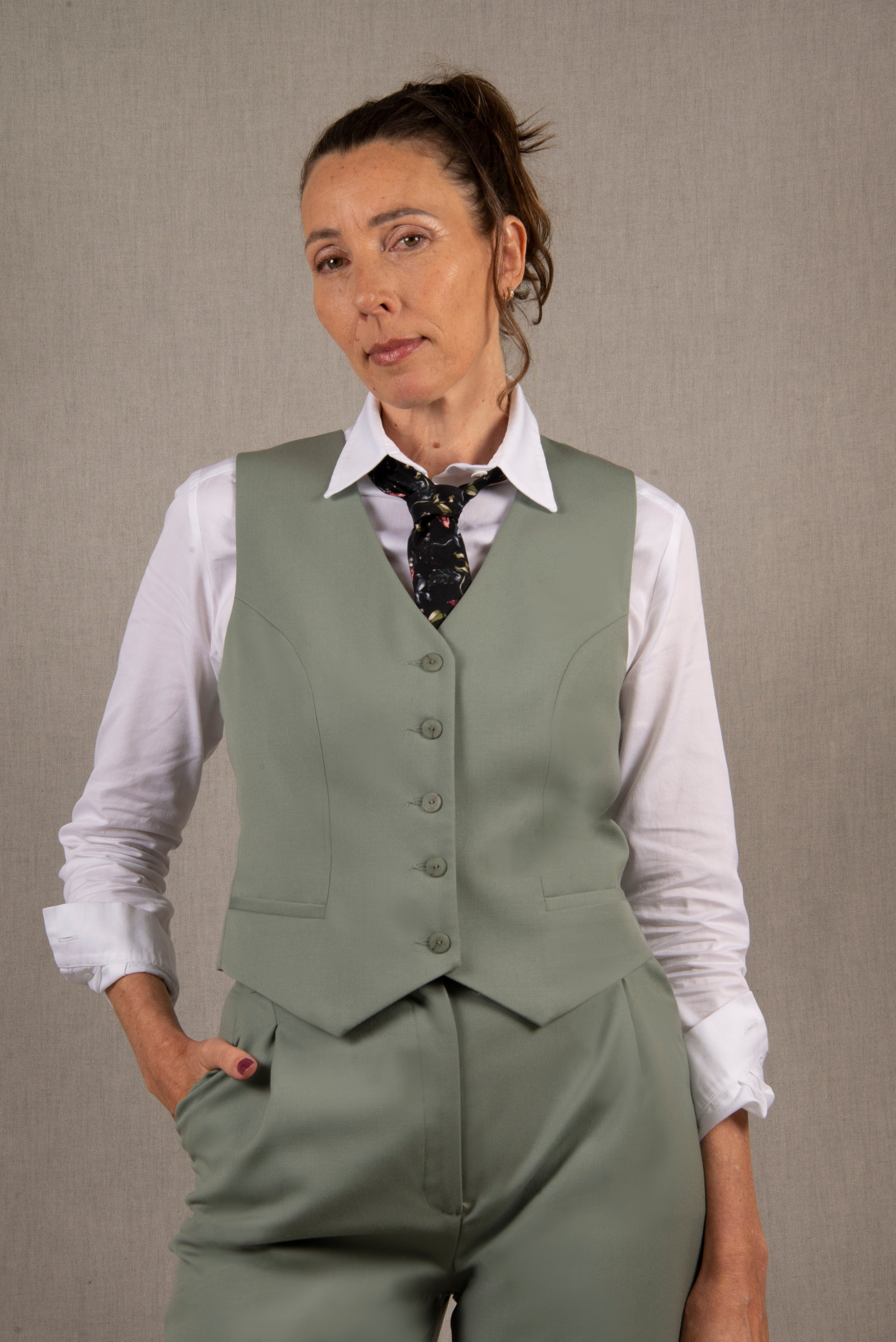 Gilet tailleur femme en 100% laine, couleur vert sauge.  Cinq boutons en nacre mates ton sur ton sur le devant, ainsi que deux poches passepoilées. La coupe de ce gilet est cintrée. La doublure est en 100% viscose avec un coloris ambré.