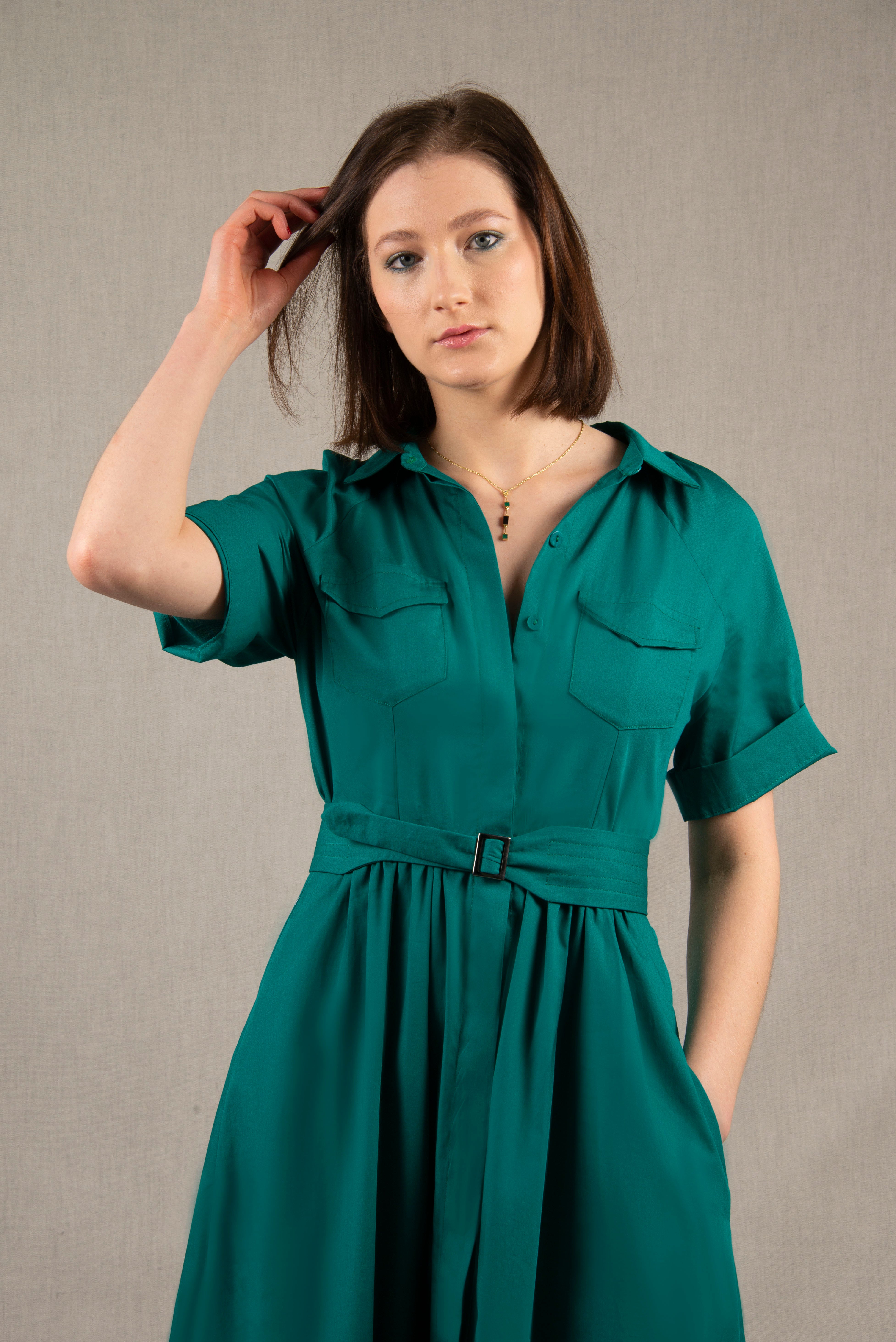 Cette robe chemisier 100% coton, à longueur sous-mollet, a un boutonnage caché sur tout le milieu devant. Elle a des manches courtes raglan avec une série de plis à la base, ainsi que deux poches plaquées à rabat sur la poitrine et deux poches sur les côtés dans la couture. Il y a un smock au milieu du dos pour cintrer la taille et une ceinture large surpiquée finie par une boucle en métal.