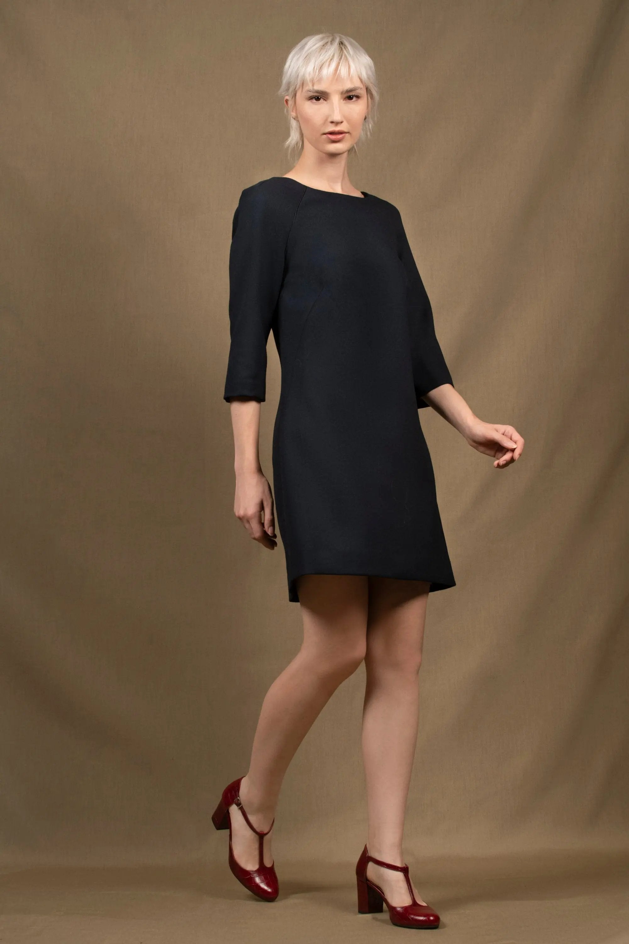 Robe droite légèrement évasée, longueur au-dessus du genou et manches trois-quarts. Col bateau bordé d’un passepoil en soie noire. Une grande pince oblique galbe le devant et deux pinces dans le dos viennent subtilement marquer la taille. Elle se ferme par une fermeture sur le côté et est entièrement doublée. Laine bleu marine.